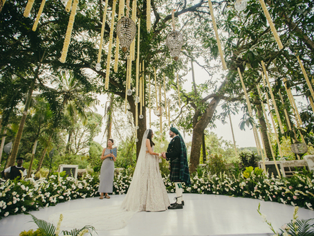 Couple exchanging vows at an outdoor destination wedding in Goa with elegant floral decor, hanging garlands, and crystal chandeliers under lush palm trees