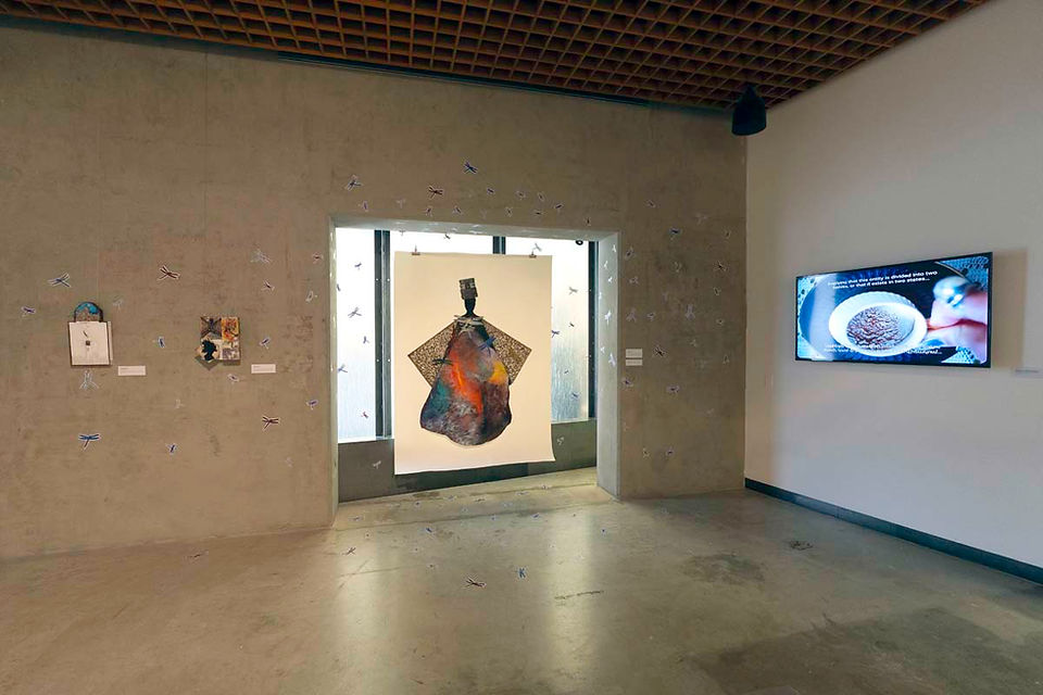 Installation view of gallery showing two perpendicular walls. Left wall features two small collage artworks, A large hanging artwork featuring a black figure of a woman with a colorful skirt, triangular wings and adorned with large dragonflies. Four small, stacked suitcases rest titled on her head. Dragonflies also adorn the wall. Other wall features a mounted tv screen showing a cup of coffee.