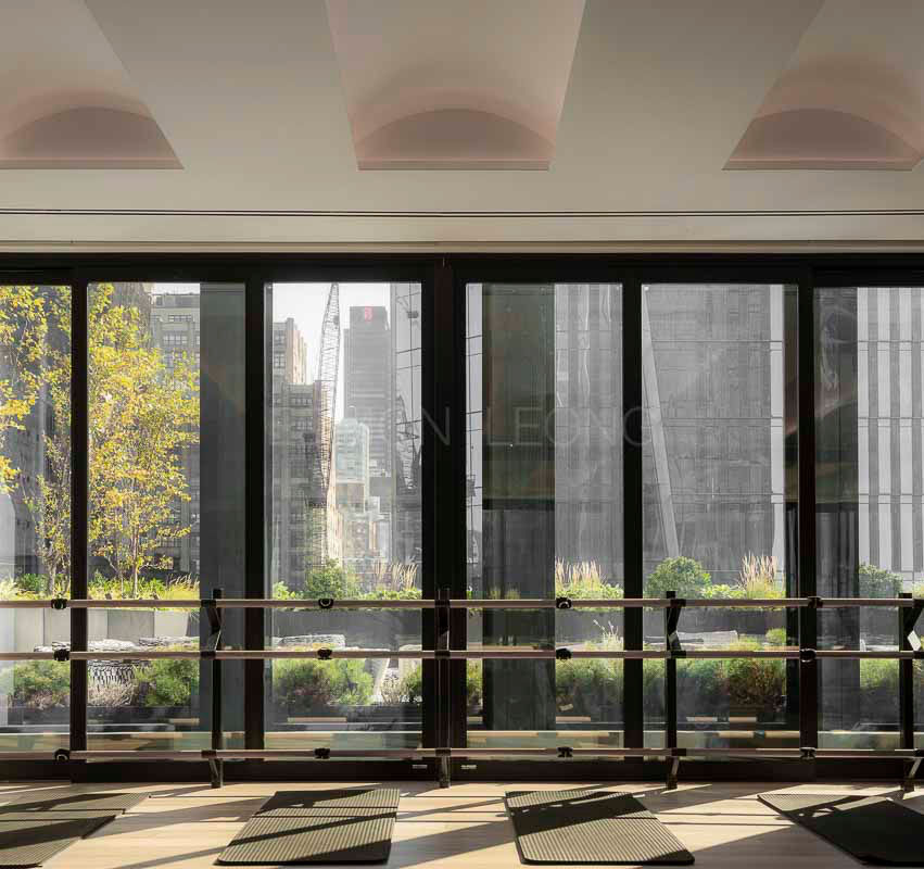 Yoga studio with mats and barre, overlooking urban skyline through large windows. Sunlit greenery outside adds a tranquil feel. High-performance luxury hospitality interior photography of Equinox Hotel New York by Edmon Leong, by Joyce Wang Studio.