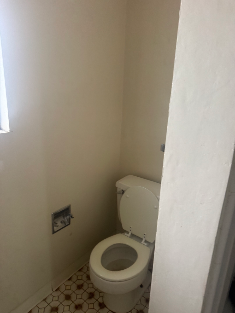 Before photo of a small Riverside bathroom toilet area with beige linoleum and old wooden shelving.