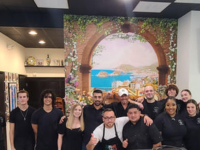 Team photo of restaurant staff and owners at Bella Vista in Fruit Cove, Florida. There are about 15 people smiling at the opening while wearing black uniforms with logo shirts, standing in front of a colorful mural of Italy inside the restaurant.