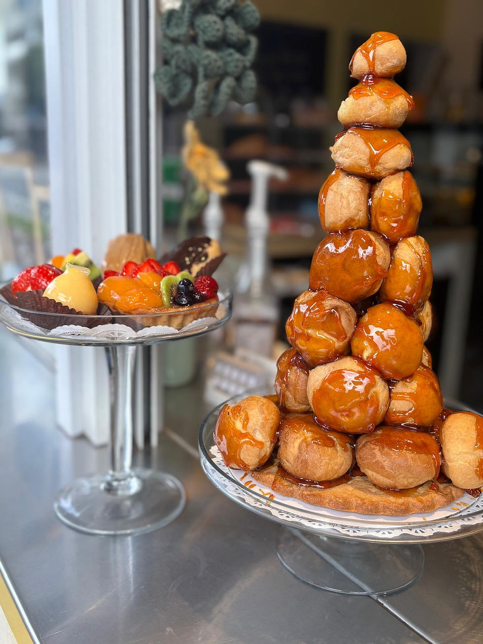 Tower of caramel-glazed pastries beside a fruit tart on a glass stand. Displayed in a bakery with a cozy background.