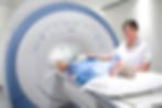 A nurse assists an older patient in a blue gown into an MRI scanner in a bright, clinical room. The nurse is smiling reassuringly.