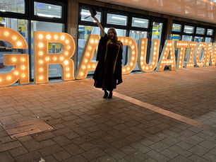 A graduate stands in front of a lit up graduation sign, holding a graduation hat above her head