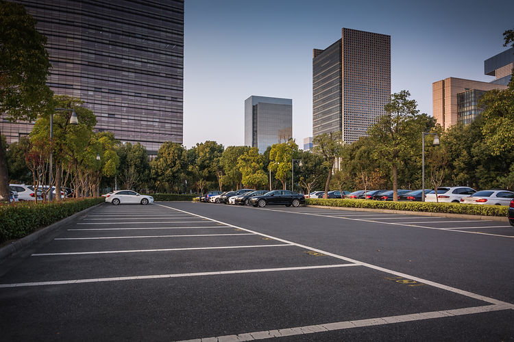 An empty parking lot with a few cars near trees and modern skyscrapers