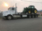 A white flatbed truck carrying a green tractor is parked on a road