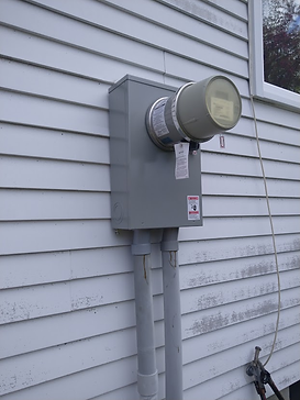 A gray electrical meter box mounted on a white wooden wall with two conduit pipes leading down