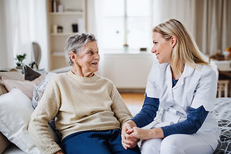 An elderly woman in a beige sweater sits holding hands with a smiling nurse