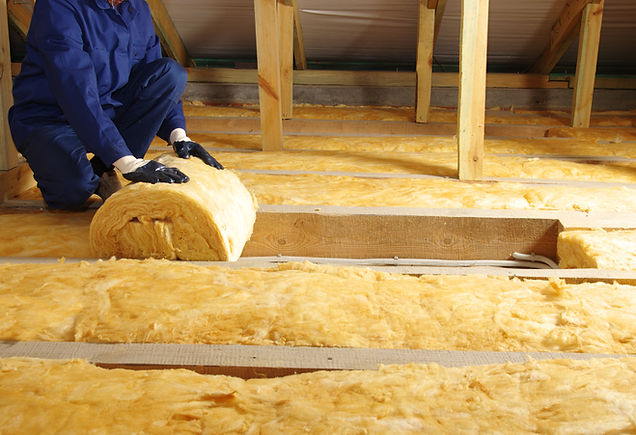 A person wearing blue overalls and gloves installs yellow insulation rolls in an attic