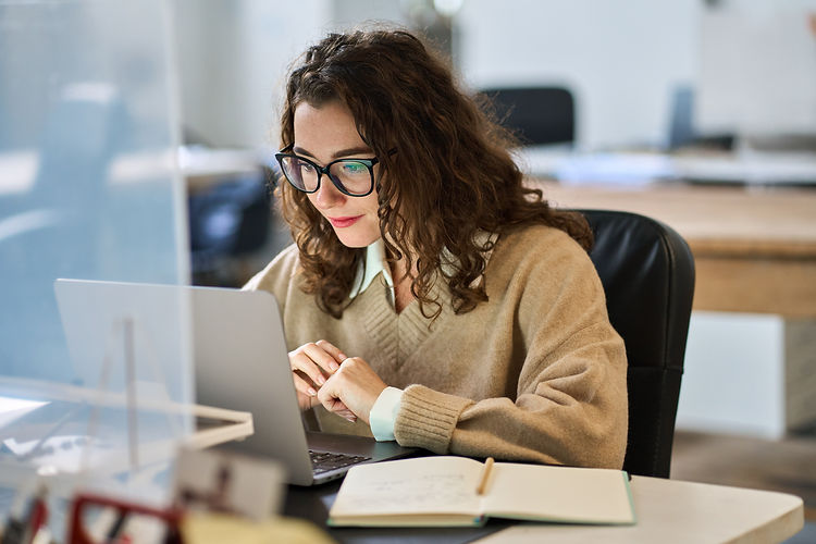 A woman with curly hair and glasses is focused on a laptop at a desk