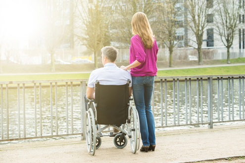 Rearview of man in a wheelchair and woman acting as a caregiver standing beside him looking out at a lake