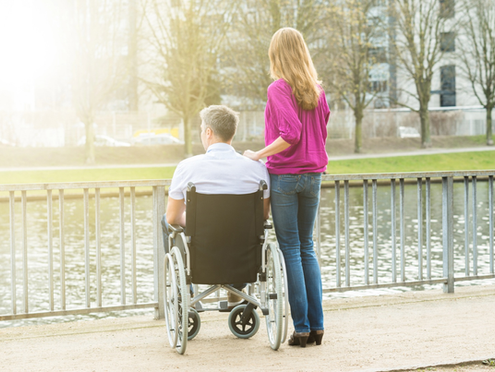 Rearview of man in a wheelchair and woman acting as a caregiver standing beside him looking out at a lake