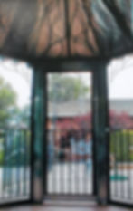 View through an ornate gazebo opening reveals a sunlit patio with iron furniture, set against a back