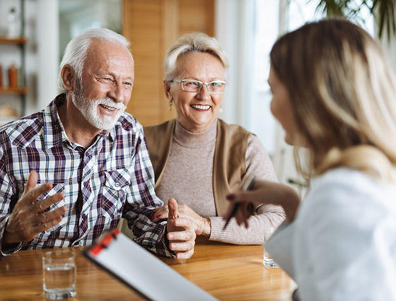 Un couple de personnes âgées, assis à une table, sourient et discutent avec une femme tenant un bloc-notes.