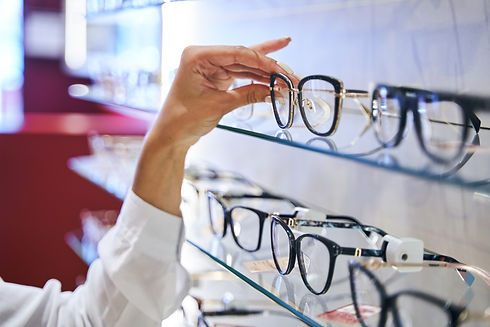 A hand reaches for black-rimmed glasses on a glass shelf lined with eyeglasses