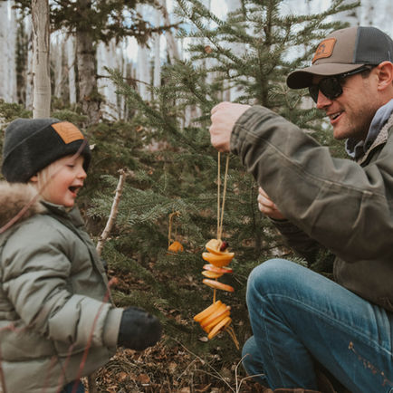 Man and child hang orange slices on small pine tree in forest. Both wear jackets and hats, smiling. Birch trees in background, autumn mood.