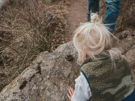 Child observing animal droppings on a large rock beside a trail. Dry grasses surround, and a person in jeans stands nearby. Neutral colors.
