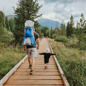 Man with blue hiking backpack carrying an infant and a toddler walk on wooden boardwalk in lush forest, mountains in background. Child has arms outstretched, joyful mood.