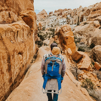 A man wearing a blue hiking backpack carrying a child in a striped sweater and brown hood hikes own a narrow red rock fin on a primitive trail in Arches National Park