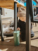 A child in a tan beanie drinks from a green tumbler on a wooden deck, surrounded by melting snow under a sunny blue sky.