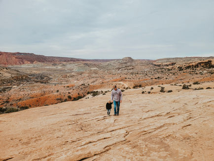 Man in grey hoodie and jeans walking on a rocky desert landscape with a small child in jeans and a black fleece. Overcast sky and vast, rugged terrain in the background.