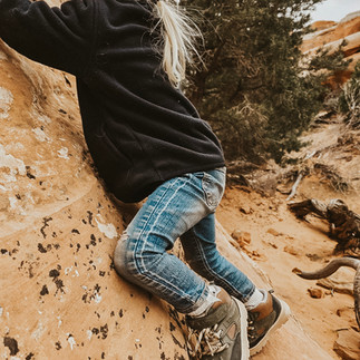 A toddler in hiking boots, jeans, and a black fleece rockclimbs up a steep section of trail in Arches National Park