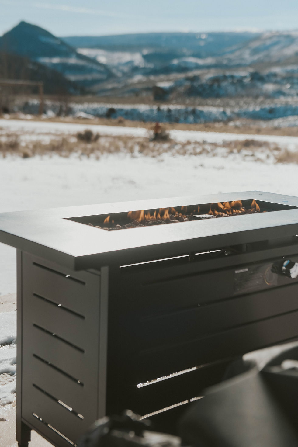 Outdoor fire pit with flames, set against a snowy mountain landscape under a clear blue sky, exuding a warm, serene atmosphere.