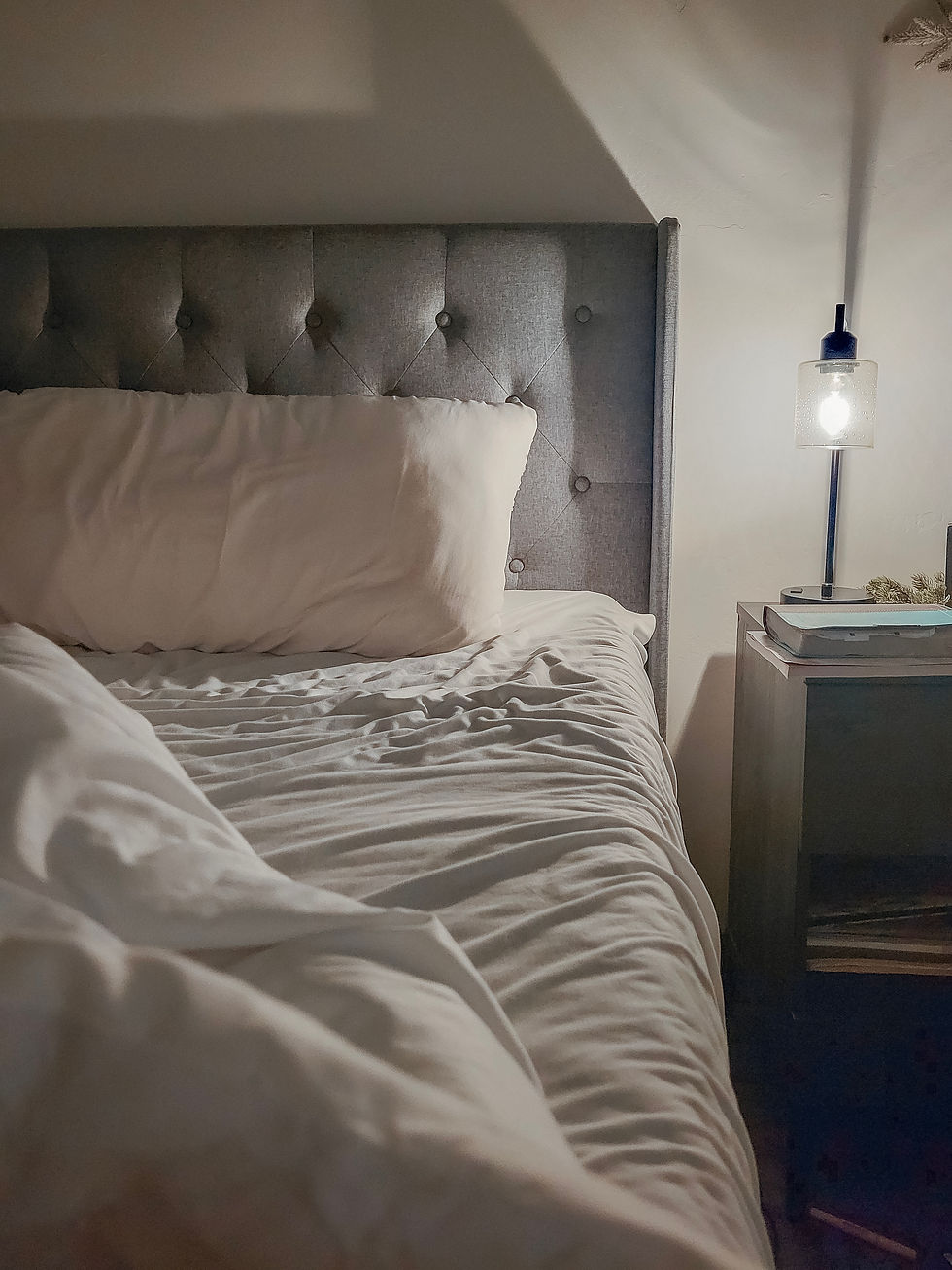An unmade bed with white linens and a gray tufted headboard. A lit lamp sits on a bedside table, creating a cozy, inviting mood.