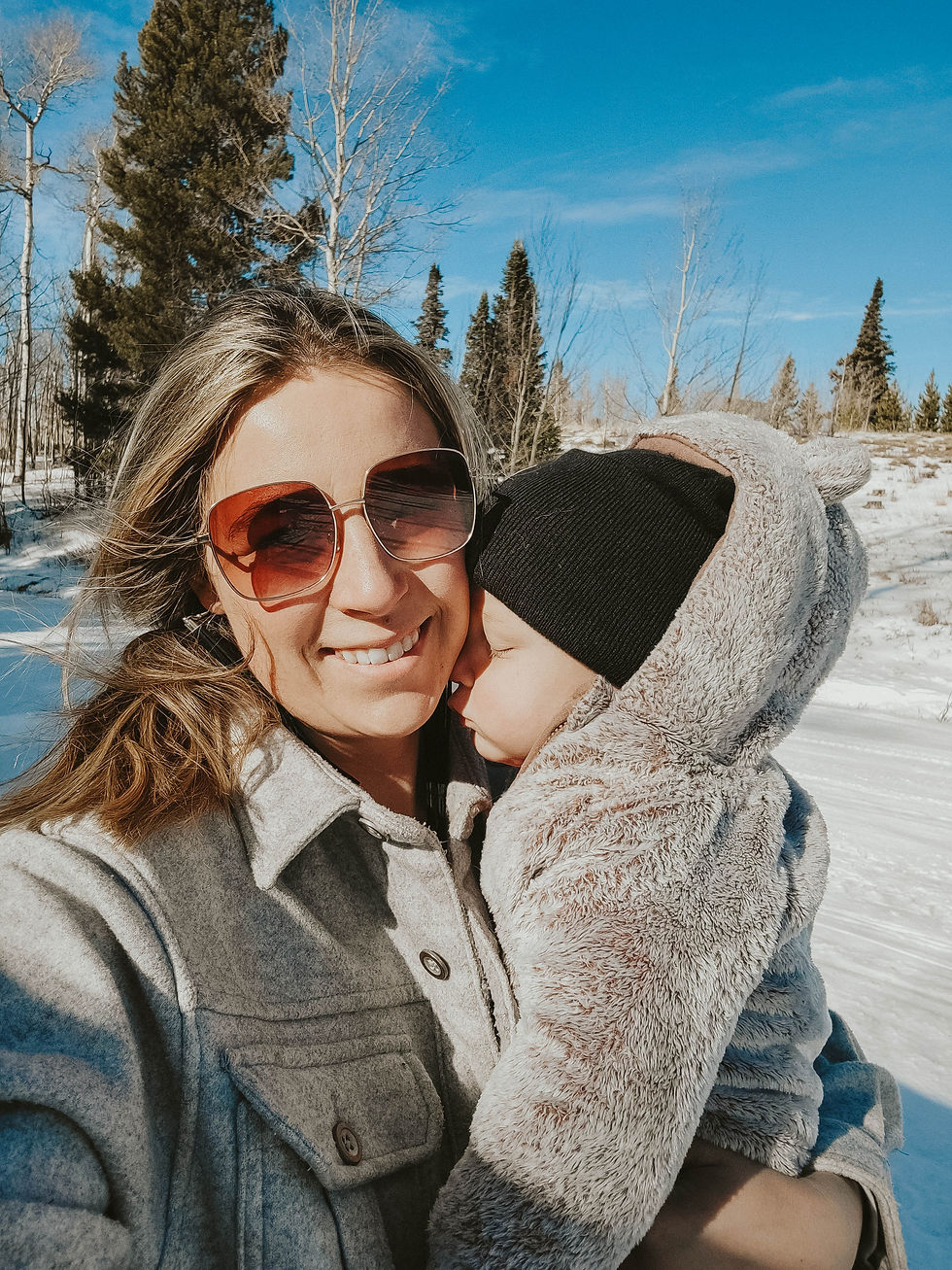 Woman in sunglasses holds a sleeping baby in a furry coat against a snowy forest backdrop. Bright blue sky, serene and joyful mood.