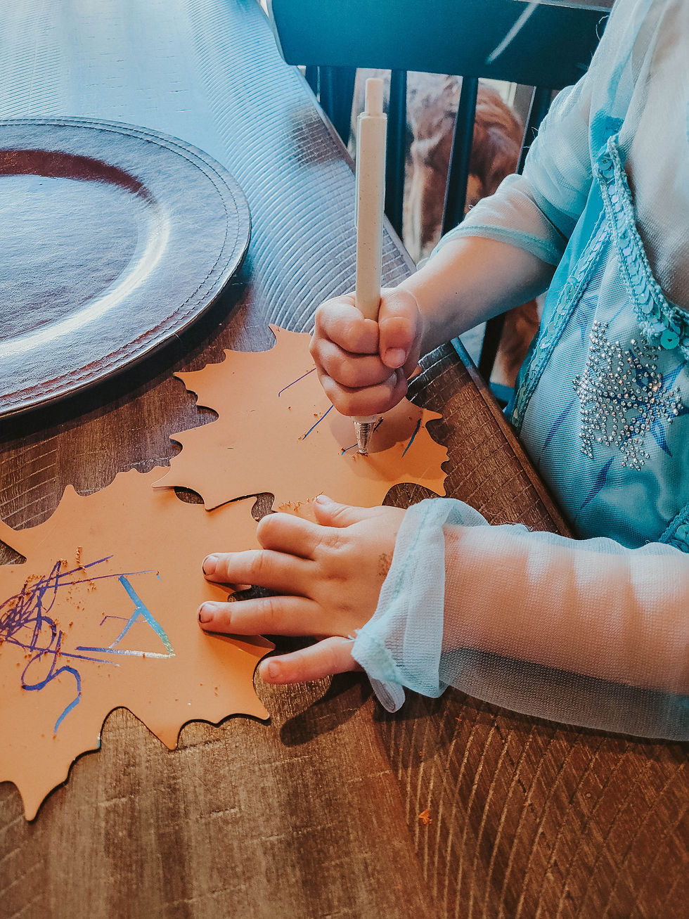 Child in blue dress draws on orange leaf-shaped paper with pencil at wooden table. Plate nearby, warm natural light.