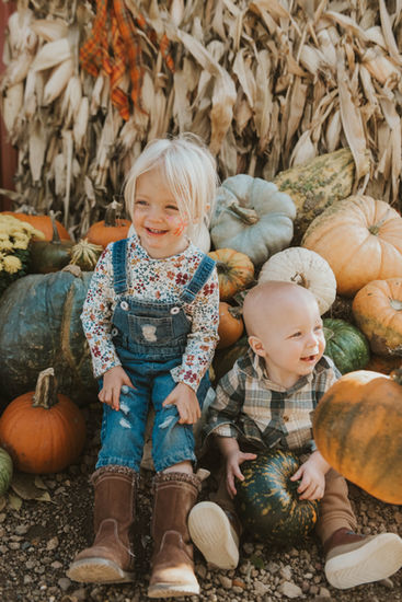 A toddler girl and infant boy sit in front of cornstalks and a pile of multi colored pumpkins, smiling in a colorful autumn scene. A sweet moment of seasonal celebrations and family adventures