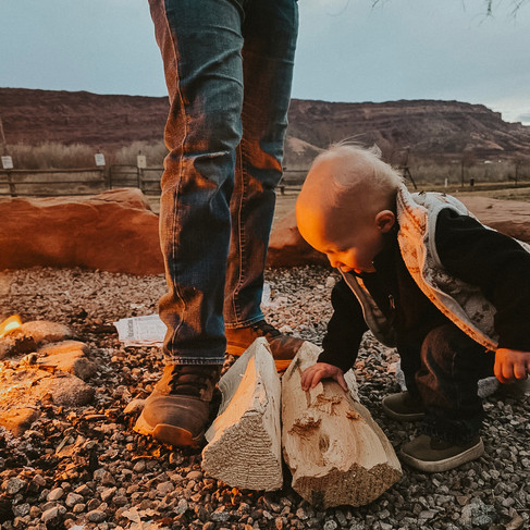 A toddler boy in a vest touches a piece of firewood beside a glowing campfire, the fire light illuminating his face and the legs of an adult in jeans with an orange glow. A desert mesa is seen in the background of this dusky scene.