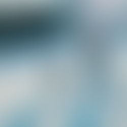 A man in jeans and a brown jacket walks behind a toddler in a green snowsuit on skis. The child is held by a ski training harness, skiing down a snowy mountain slope under a sunny blue sky. 