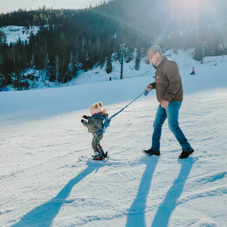 A man in jeans and a brown jacket walks behind a toddler in a green snowsuit on skis. The child is held by a ski training harness, skiing down a snowy mountain slope under a sunny blue sky. 