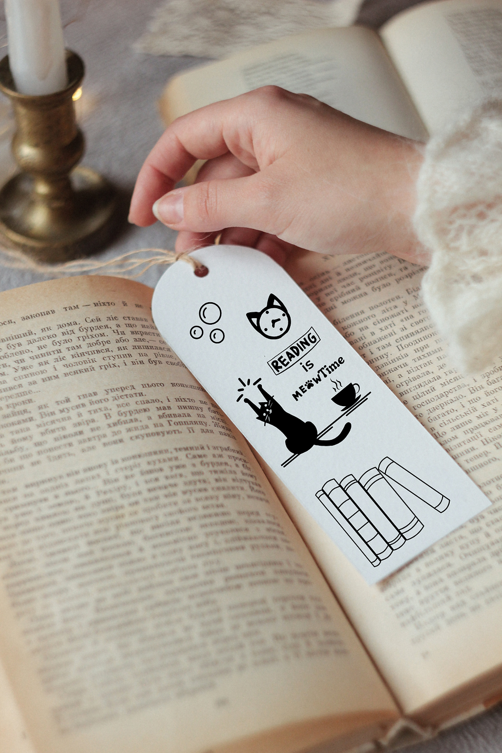 A hand holds a cat bookmark in an open book. The bookmark shows a cat, books, and text: "Reading is Meowtime." A candle is nearby. Cozy setting.