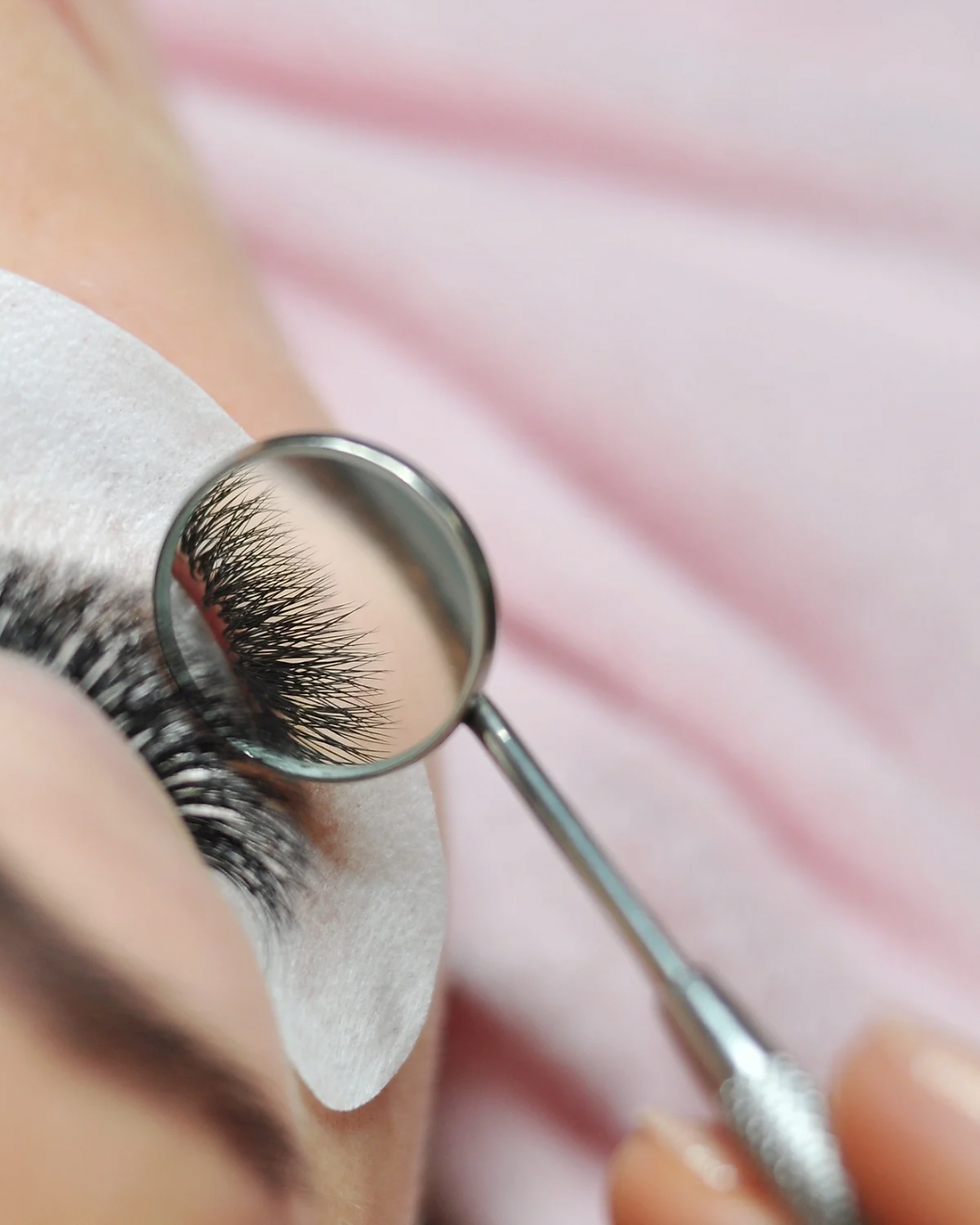 Close-up of eyelashes being examined with a round mirror. Eye patch underneath. Background is soft pink, conveying a serene mood.