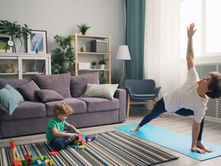 Child plays with blocks on a striped rug while an adult practices yoga nearby in a cozy living room with a purple sofa and plants.
