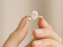 Fingers hold a gold ring with a large oval diamond and smaller stones, set against a soft beige background. The scene is calm and elegant.