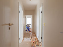 Workers in a hallway install wooden trim. White walls, doors on either side, and tools scattered on the floor evoke a busy renovation scene.