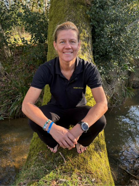 Smiling picture of Co-Founder Sam Kinsey-Briggs squatting on a mossy tree trunk by a stream. Wearing a black polo shirt, blue bracelets, and watch. Sunlit, wooded background.