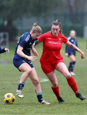 Two female soccer players compete for the ball on a grassy field. One wears red, the other navy blue. Three more players are in the background.
