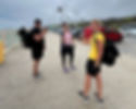 Three people with backpacks hold trekking poles, smiling on a seaside promenade. Overcast sky, red van, and parked cars in the background.