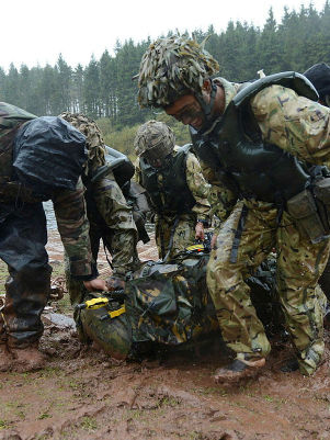 Soldiers in camouflage gear carry equipment through mud by a lake, with a forest in the background. The scene is rainy and intense.