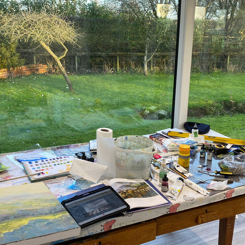 Garden room artist studio, a desk that someone has been working at with paints, canvas and brushes . The garden room has a large window that looks out onto the garden.