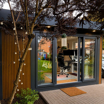 A 5.8m x 5.6m bespoke  garden room with vertical slat cladding on the front. It has patio in front of it with a tree with fairy lights to the left. 