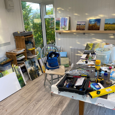Garden room artist studiointerior with a table and paints and many paintings. There is also a corner window looking onto countryside.