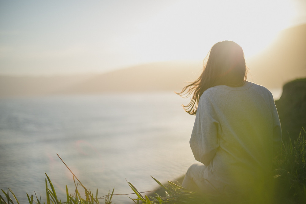 Woman sitting on the grass by the ocean who is staying in the present moment while on vacation