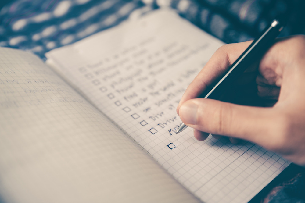 A travel budget checklist in a notebook and a woman's hand adding to it with a black pen.