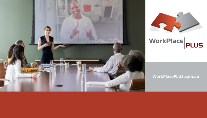 Small team of professionals at board room table. Woman standing making a presentation. Another woman smiling via video conferencing.