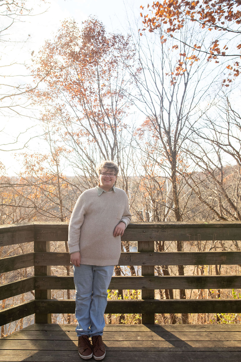 Person in a beige sweater and jeans leans on a wooden railing. Autumn trees with orange leaves in the background. Bright, sunny day. Charleston High School senior photo Doudna Fine Arts Center.